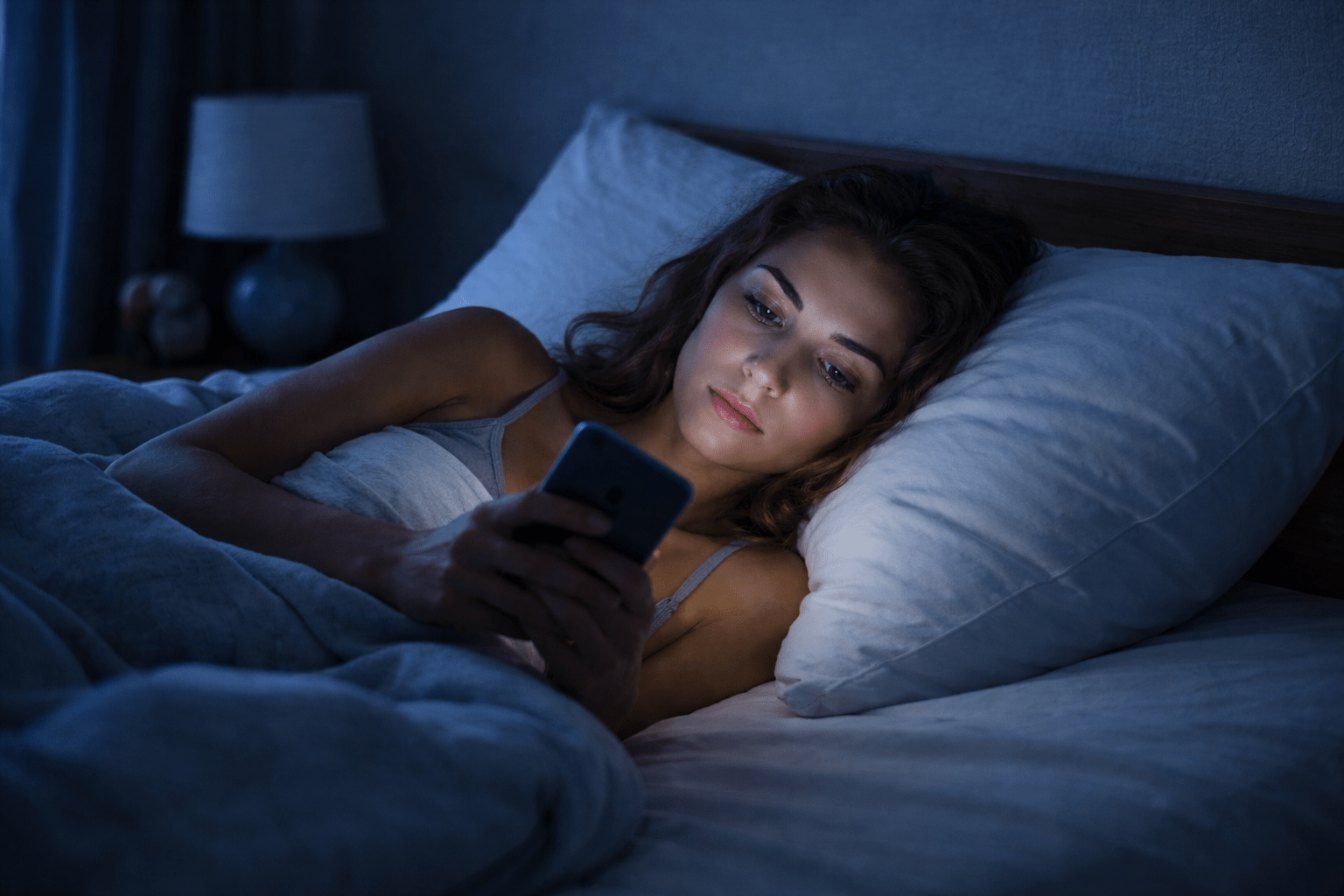 Persona acostada en la cama mirando el celular, con el rostro iluminado por la luz azul de la pantalla en una habitación oscura. La escena refleja estimulación nocturna, dificultad para desconectar y alteración del descanso por el uso de redes sociales.