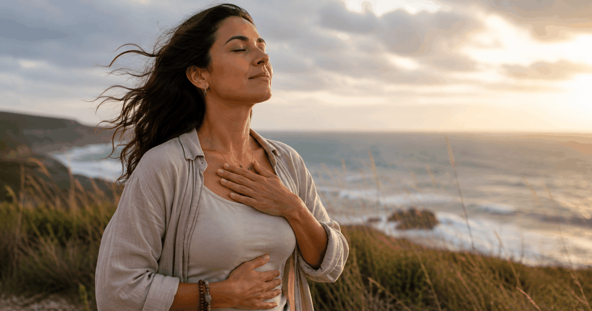 Mujer latinoamericana respirando profundamente frente al mar al atardecer, con una mano en el pecho y otra en el abdomen, en medio del viento.