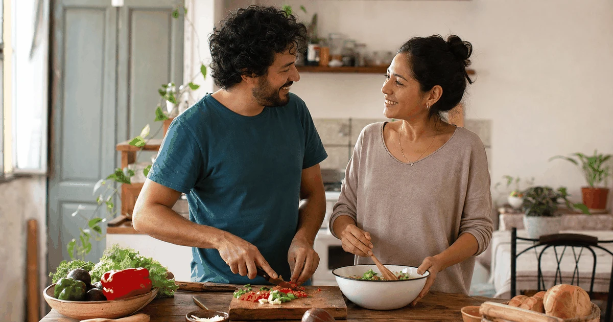 Pareja adulta latinoamericana prepara una comida y conversa con calidez en una cocina, representando cooperación, reciprocidad y conexión cotidiana.